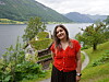 Kvinne i rød bluse står ved en sti med fjord, fjell og grønn natur i bakgrunnen.