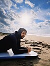 Person i svart våtdrakt ligger på et surfbrett på stranden under en lys himmel.