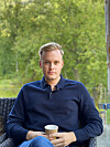 En mann sitter i en stol ute og holder en kaffekopp foran seg, med grønne trær i bakgrunnen.